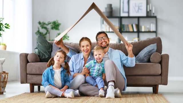 Das eigene Zuhause bedeutet auch Sicherheit für Kind und Kegel junge Familie sitzt in ihrem Wohnzimmer und hält ein Modell eines Hausdaches aus Pappmaché über den Köpfen