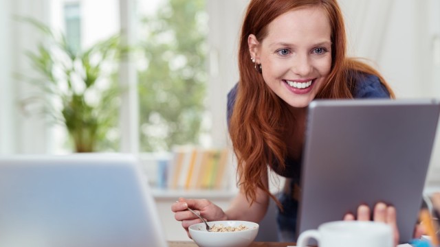 Frau isst Müsli und schaut dabei strahlend auf ihren Computer Monitor
