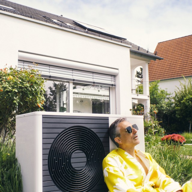 Ein Mann lehnt sich an seine Wärmepumpe im Garten, deren Installation er von der Steuer absetzen kann Ein Mann lehnt sich gegen seine neu erworbene Wärmepumpe im Garten