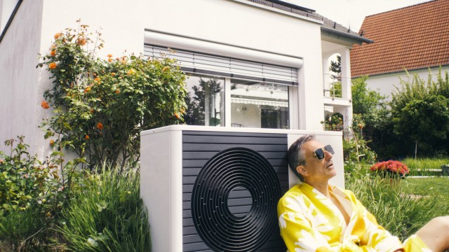 Ein Mann lehnt sich gegen seine neu erworbene Wärmepumpe im Garten Ein Mann lehnt sich gegen seine neu erworbene Wärmepumpe im Garten